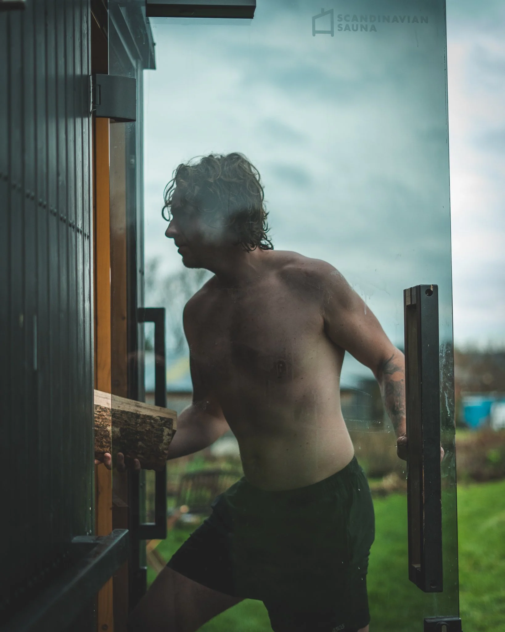 At Unfold Sauna Club A shirtless man with curly hair opening a glass door with a wooden handle, outside in a grassy area on a cloudy day.