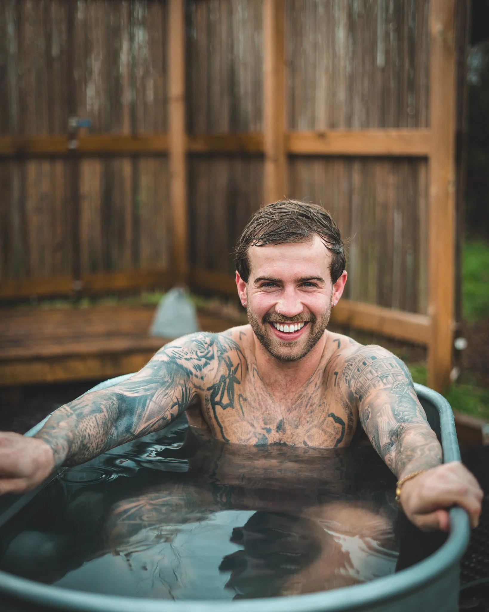 At Unfold Sauna Club A smiling man with tattoos on his arms and chest relaxing in an outdoor hot tub, holding onto the side with his arms outstretched. Wooden fencing surrounds the hot tub area.