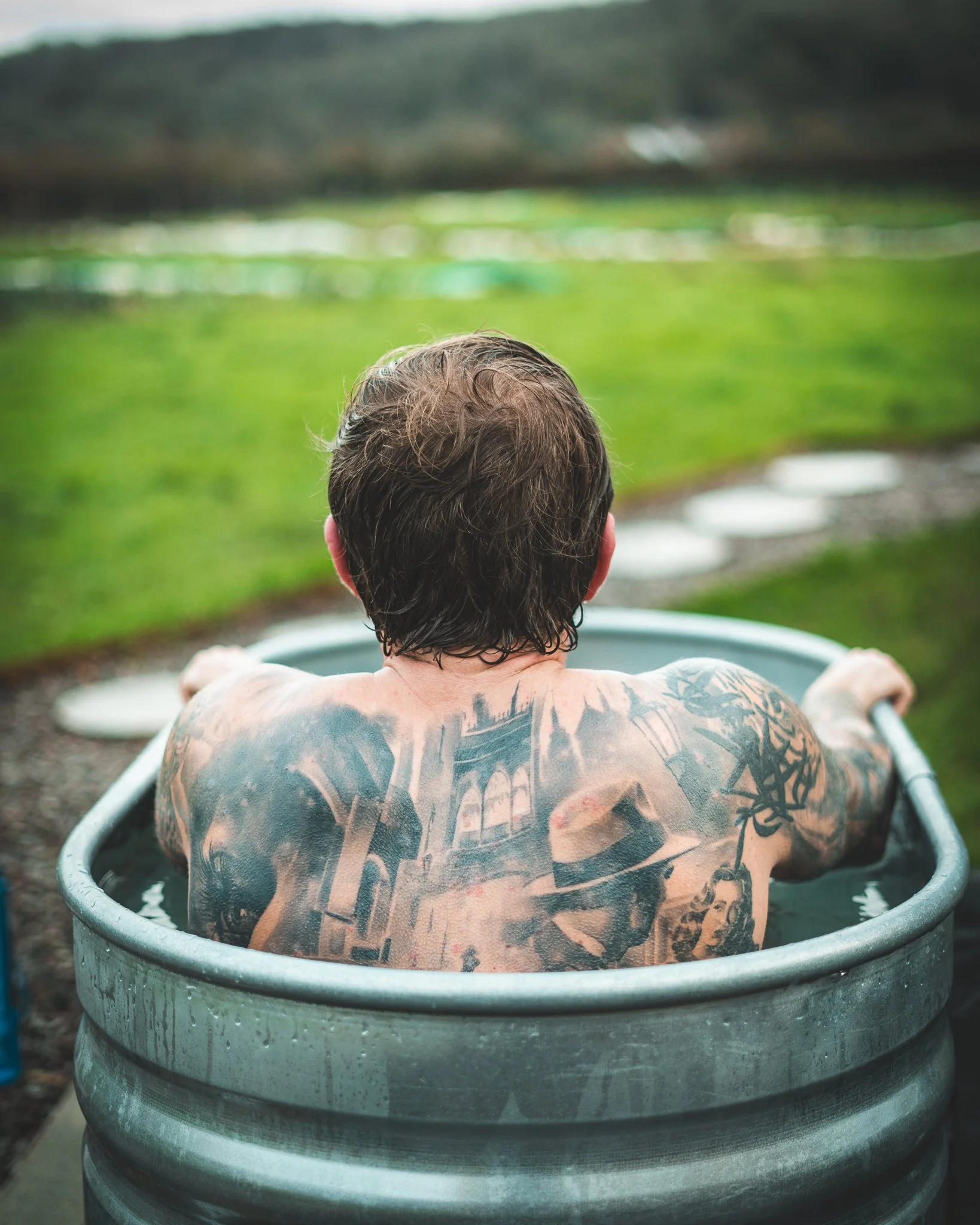 At Unfold Sauna Club Man sitting in a metal tub filled with water, with tattoos on his back, facing away in a lush green outdoor setting.