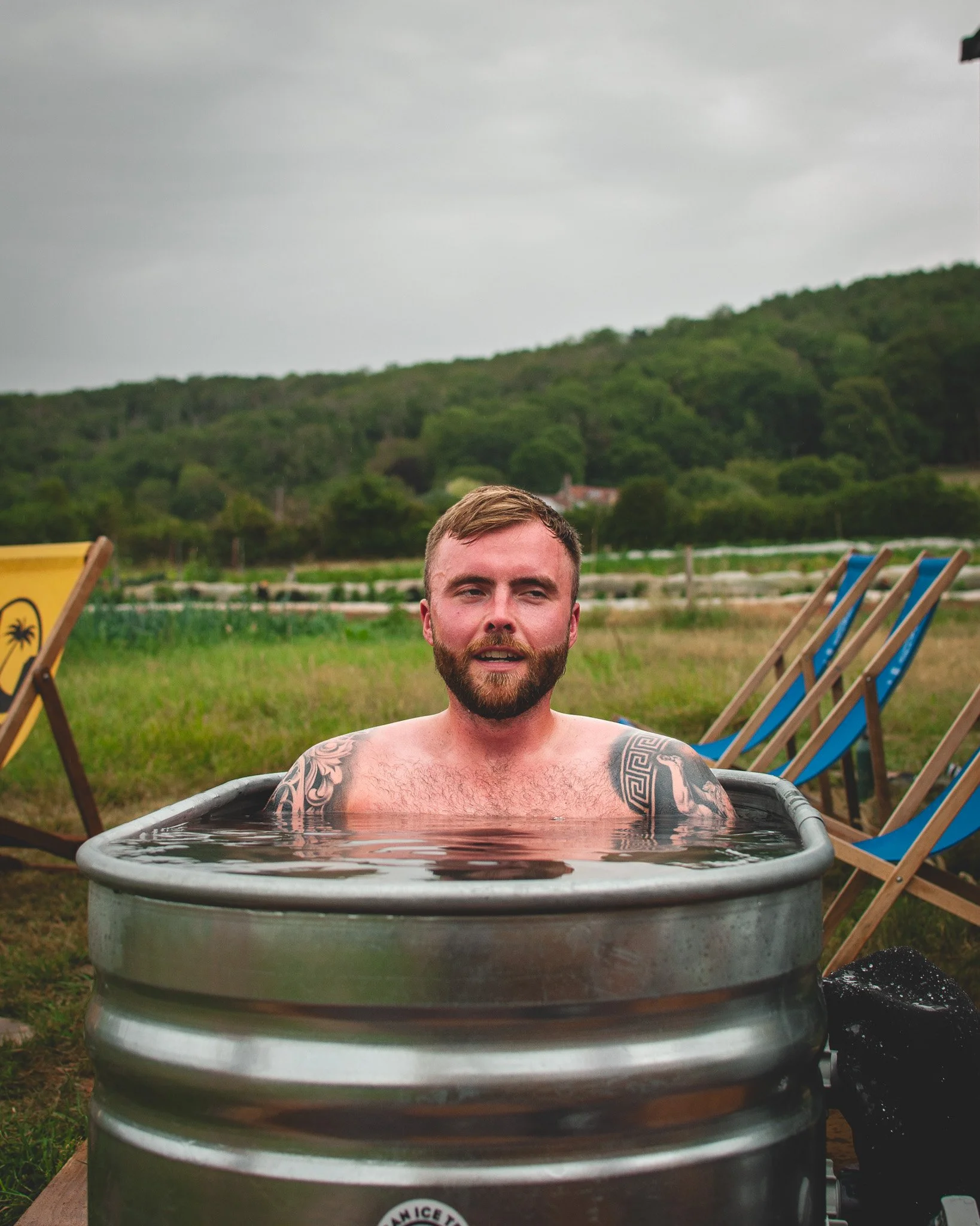 At Unfold Sauna ClubA man with a beard and tattoos relaxing in a metal tub outdoors on a cloudy day, with green hills and blue lounge chairs in the background.