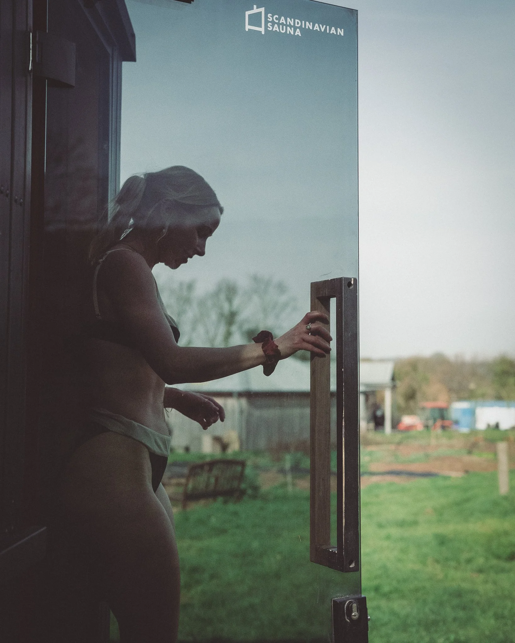 At Unfold Sauna Club A woman standing inside a sauna, with her hand on the glass door handle, looking downward. Outside the glass door, a green outdoor area and some buildings are visible. The sign above the door reads 'Scandinavian Sauna'.