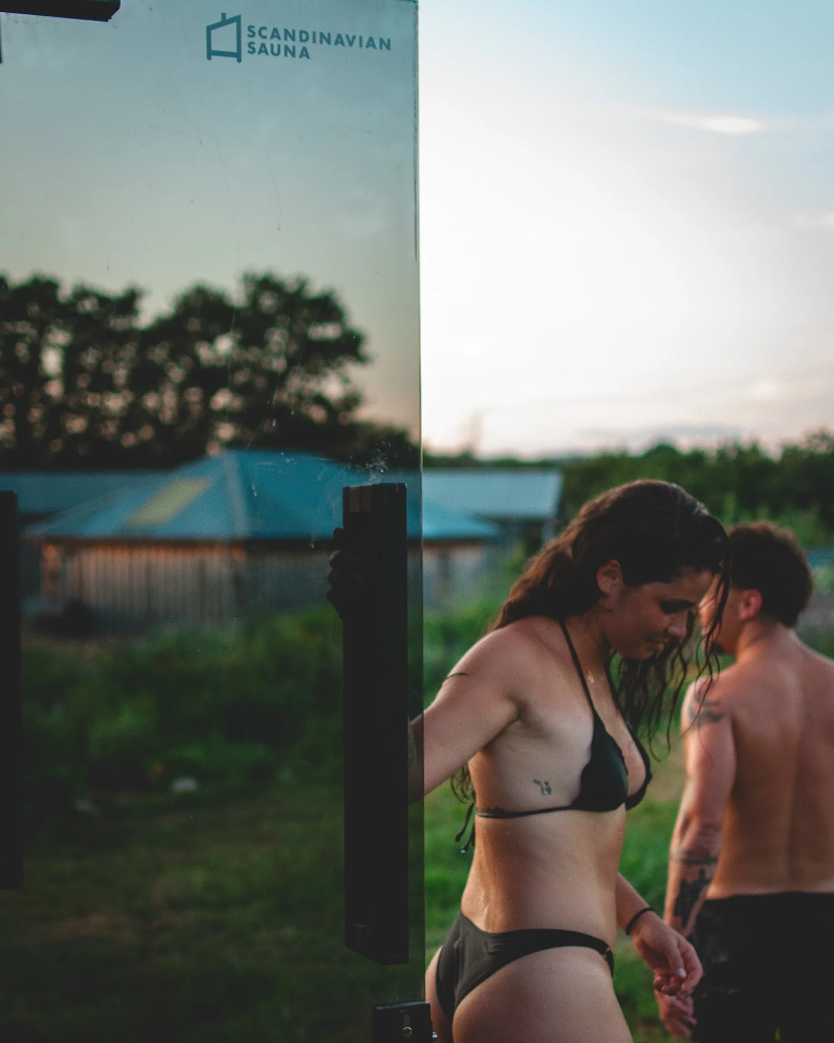 At Unfold Sauna Club Two people in swimsuits, a woman and a man, standing outdoors next to a glass panel with the logo 'Scandinavian Sauna' on it, during sunset with trees and a building in the background.