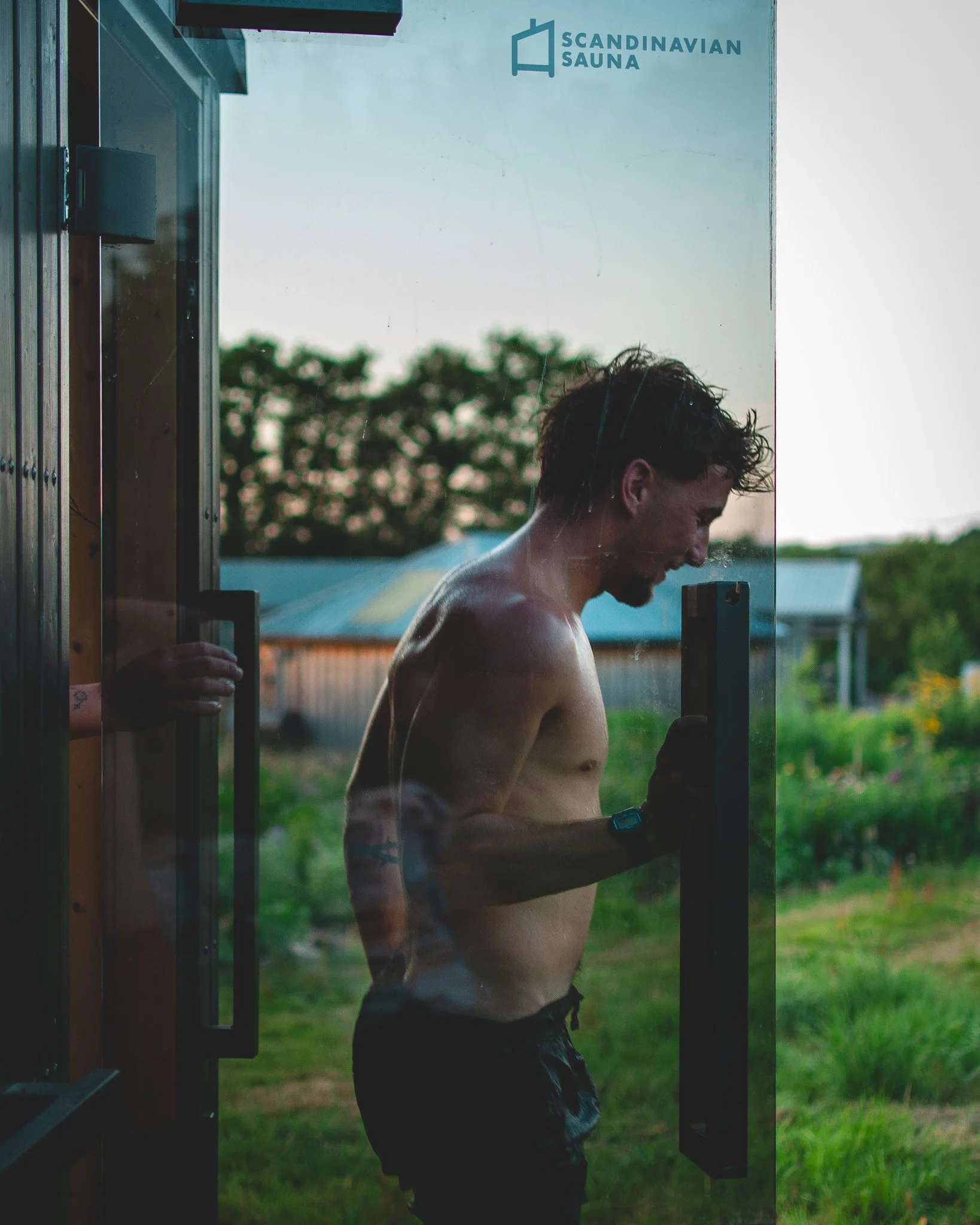 At Unfold Sauna Club A shirtless man with wet hair standing outside a sauna, looking at his phone, with a green outdoor landscape in the background.