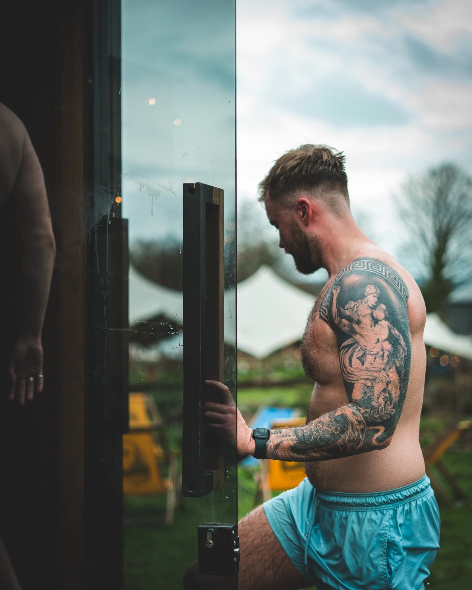 At Unfold Sauna Club A shirtless man with tattoos on his arm, wearing blue shorts and a watch, stands outside next to a glass door, looking at his reflection.