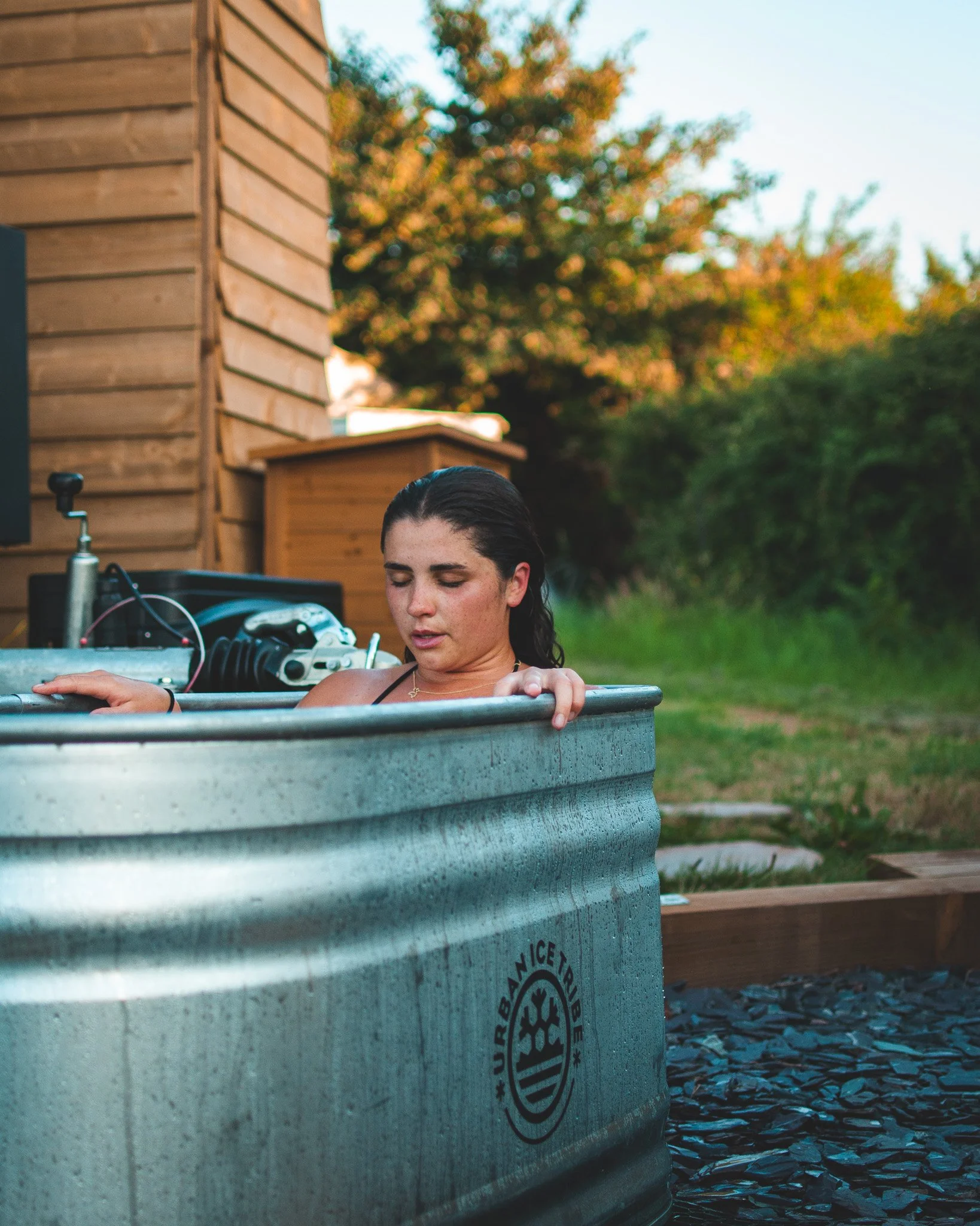 At Unfold Sauna Club A woman with dark hair in a metal tub outdoors, with electronic equipment nearby, during sunset with trees and a wooden building in the background.