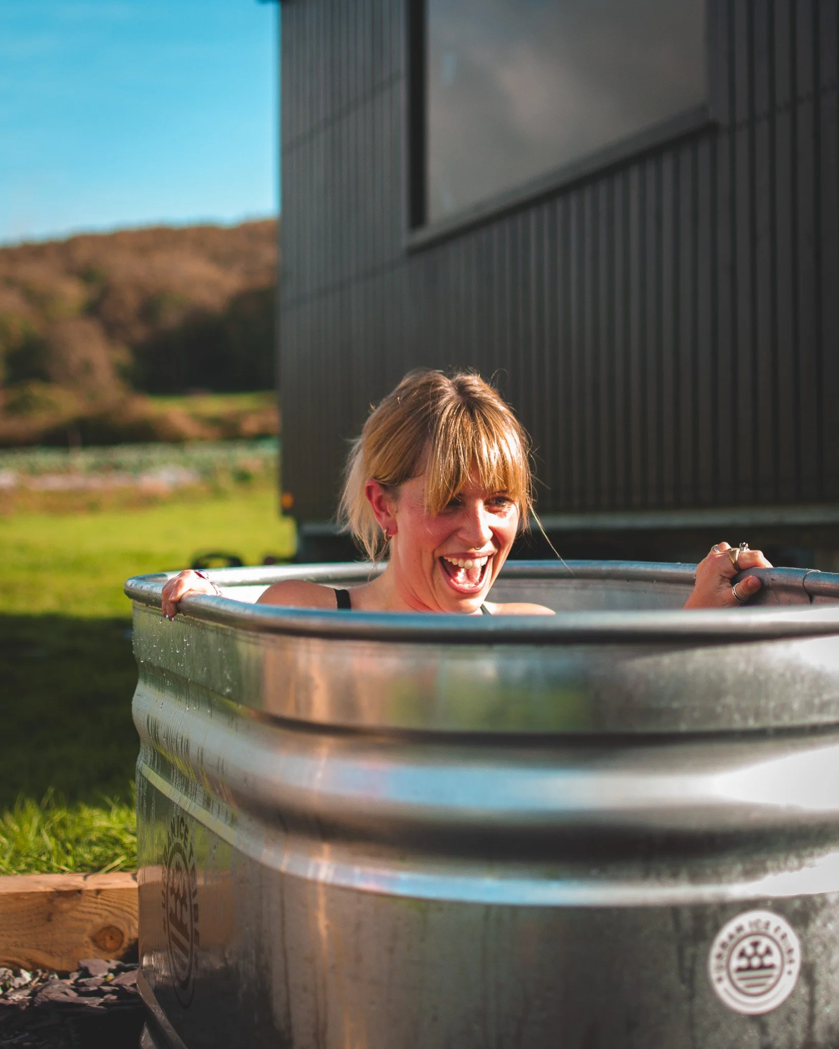 At Unfold Sauna Club A woman with blonde hair and a black tank top laughing while sitting in a metal tub outdoors during daytime.
