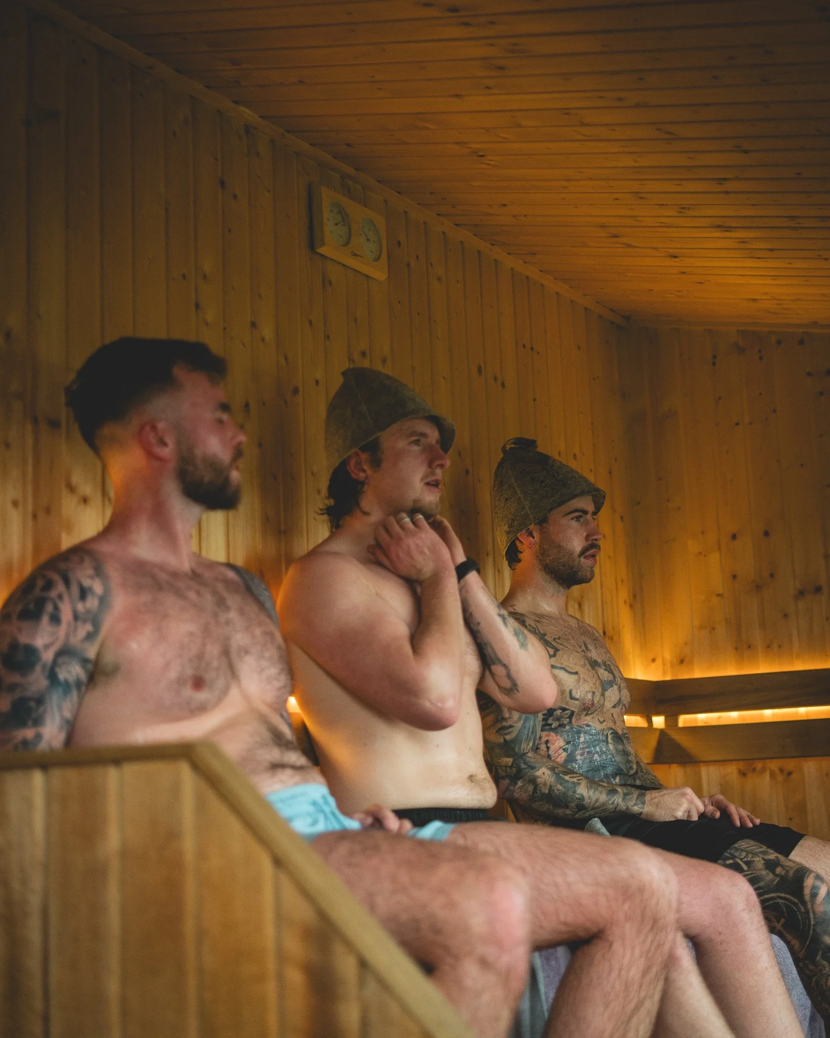 At Unfold Sauna Club Three shirtless men with tattoos sit in a sauna, wearing towels on their heads, with a wooden interior and a thermometer on the wall.