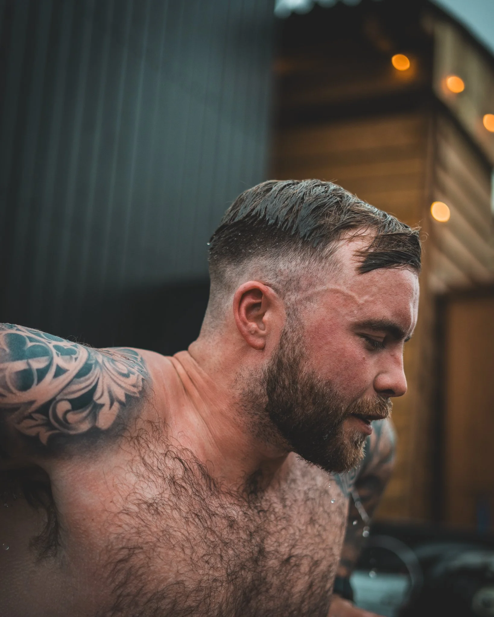 At Unfold Sauna Club A shirtless man with tattoos on his shoulder and chest, wet hair styled back, and a beard, appears to be outdoors at sunset or evening, with a modern wooden and metal structure in the background and warm lighting.