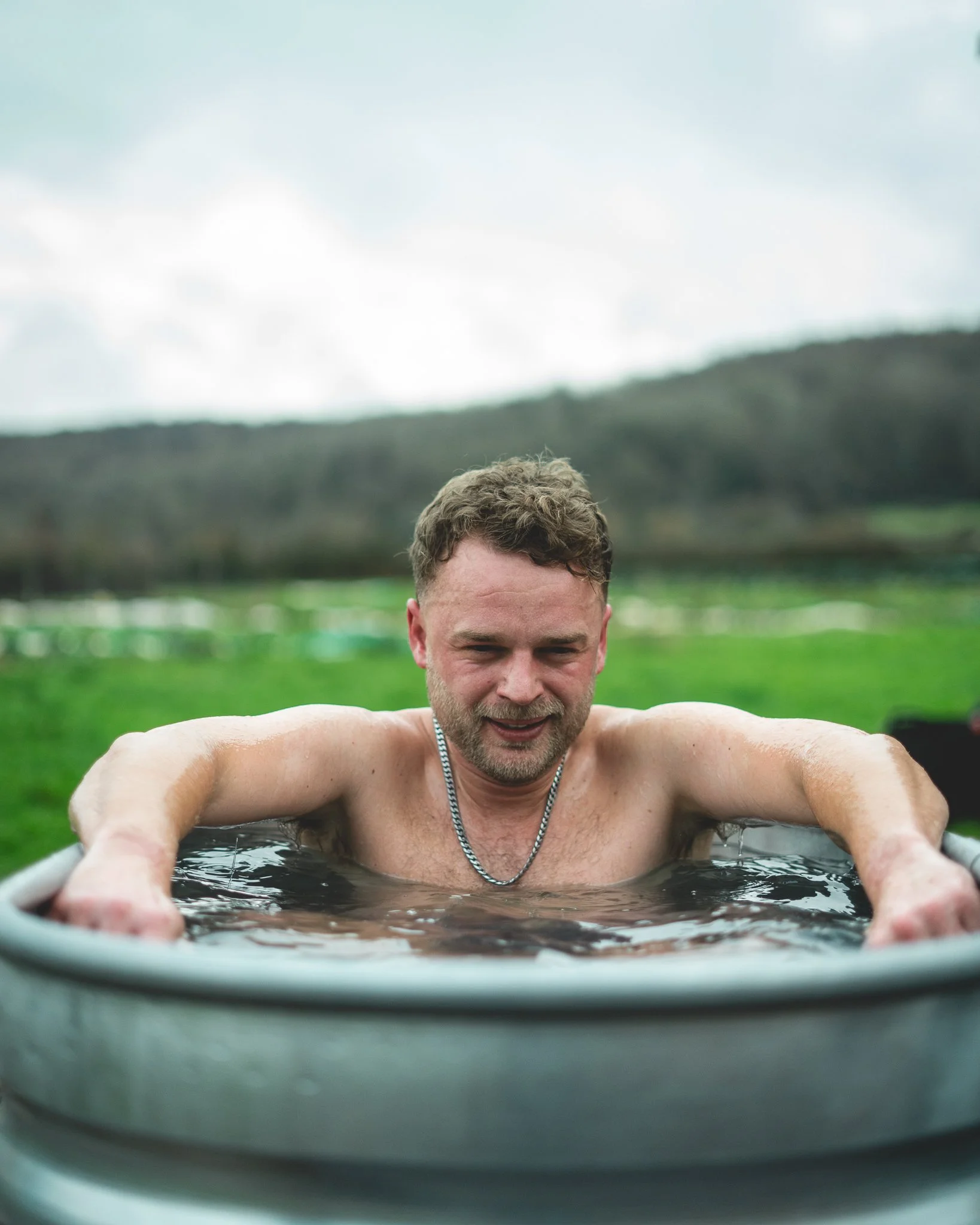 At Unfold Sauna Club A man with short hair and a beard, wearing a chain necklace, is relaxing in an outdoor hot tub or pool, with a scenic green landscape and hills in the background.