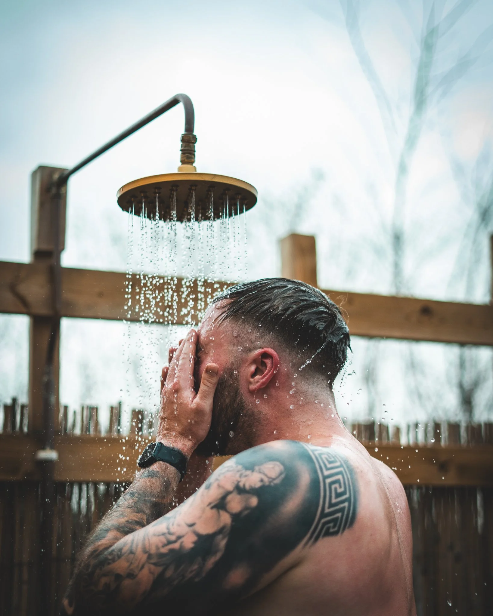 At Unfold Sauna Club Man with tattoos rinsing his face under an outdoor shower with water falling from a showerhead.