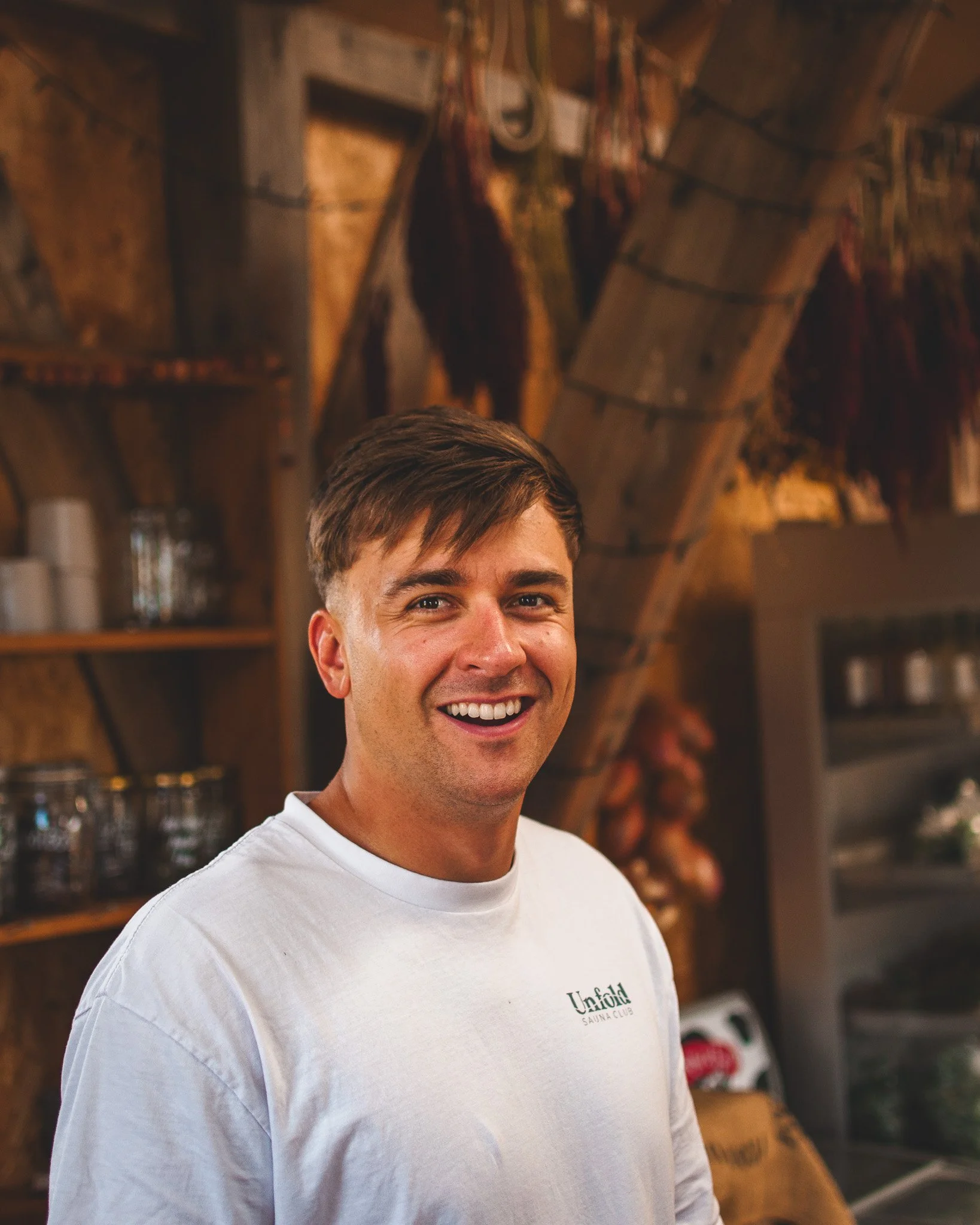At Unfold Sauna Club A smiling man wearing a white shirt standing in a rustic setting with wooden shelves and hanging dried herbs or vegetables in the background.
