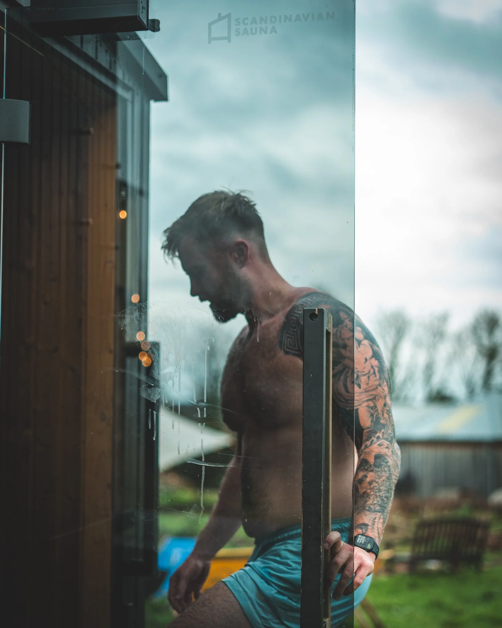 At Unfold Sauna Club A shirtless man with tattoos is looking down inside a sauna, standing by the glass door with the 'Scandinavian Sauna' logo, during cloudy weather.