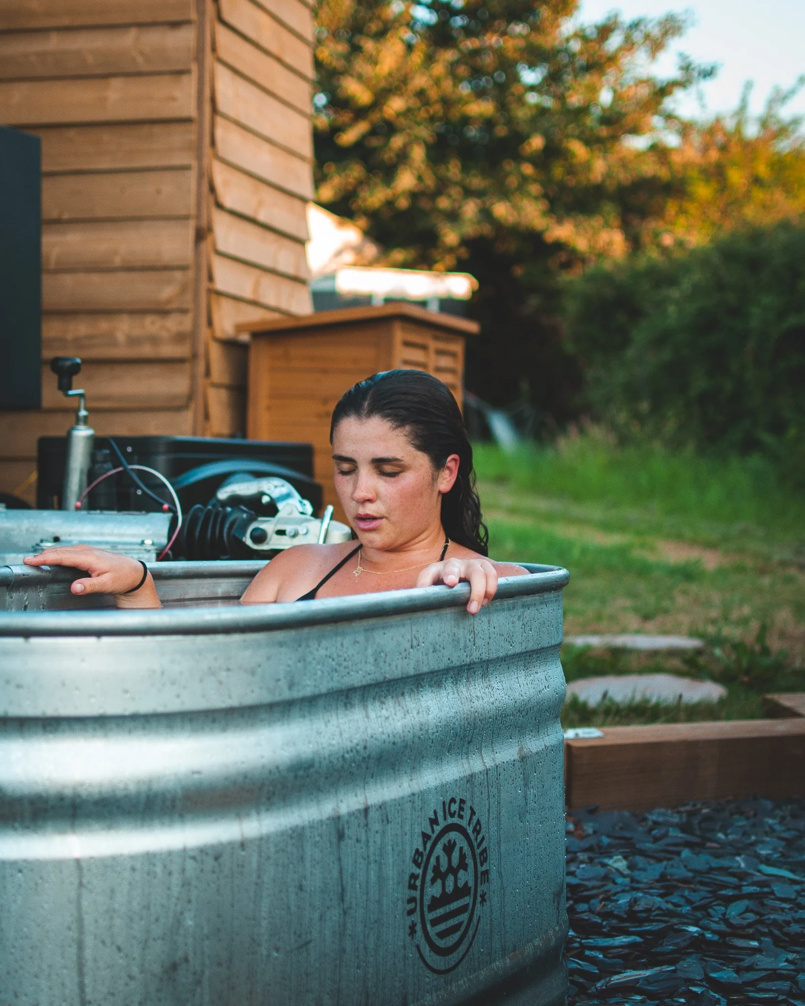 At Unfold Sauna Club Woman relaxing in a metal outdoor hot tub outdoors with a wooden shed and greenery in the background during dusk.