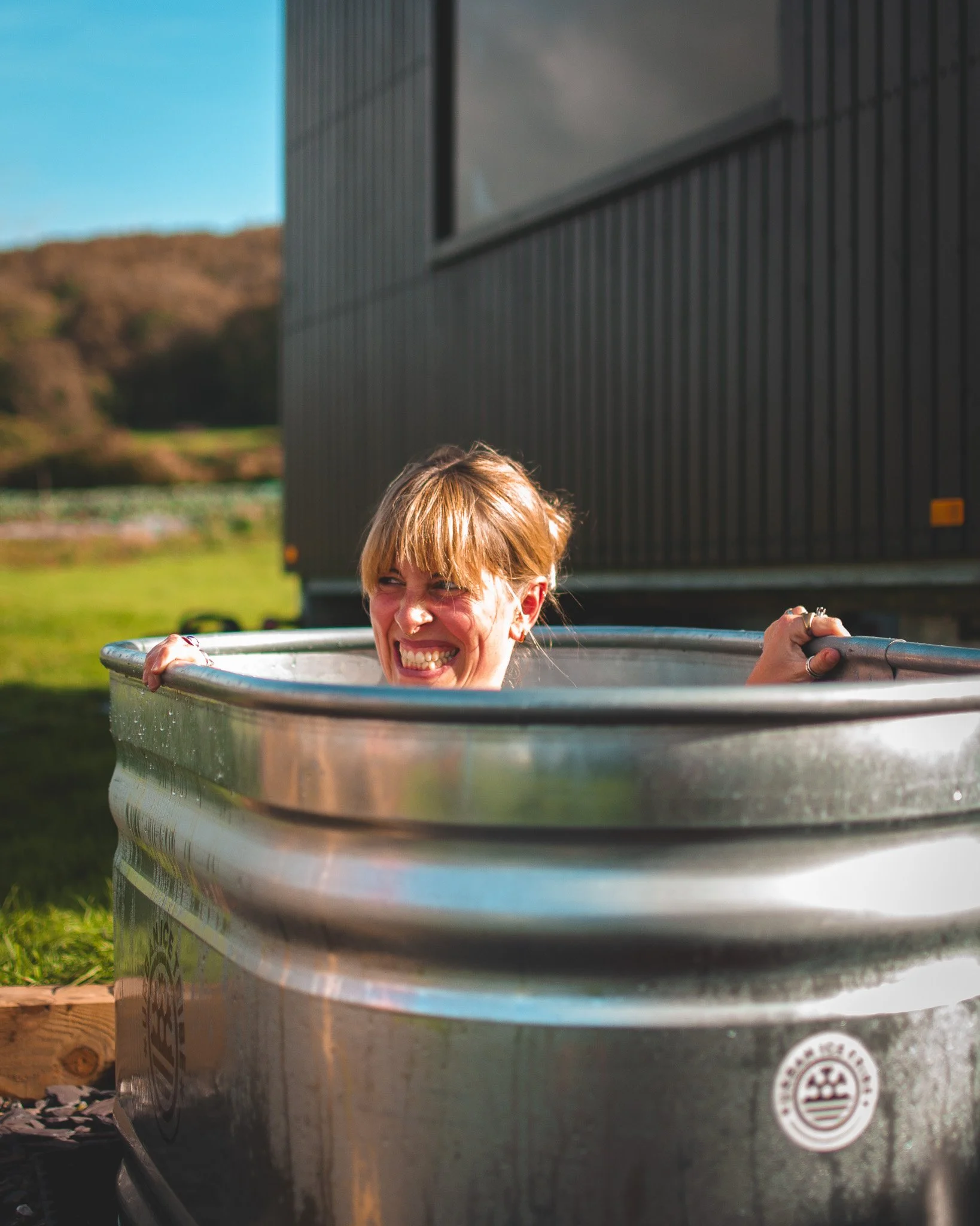At Unfold Sauna Club Woman smiling and holding onto the edge of a large metal tub outdoors with a building and green landscape in the background.