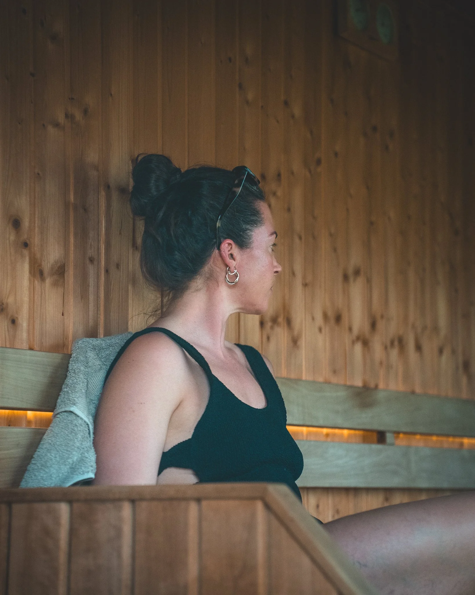 At Unfold Sauna Club A woman with dark, pulled-back hair sitting on a wooden bench inside a room with wood-paneled walls, looking to the right.