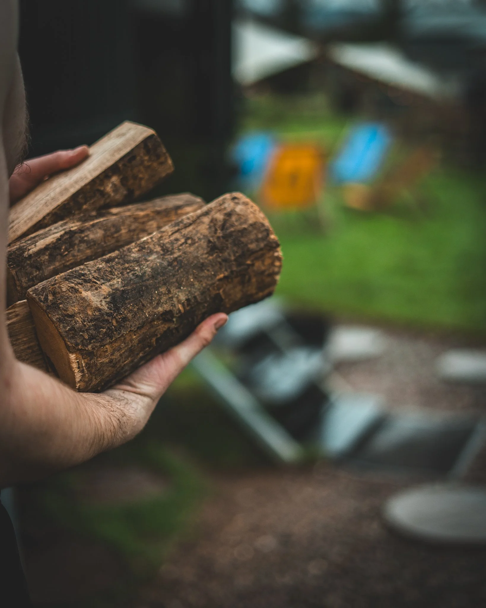 At Unfold Sauna Club A person holding a bundle of firewood outdoors, with blurred background including an umbrella and chairs.
