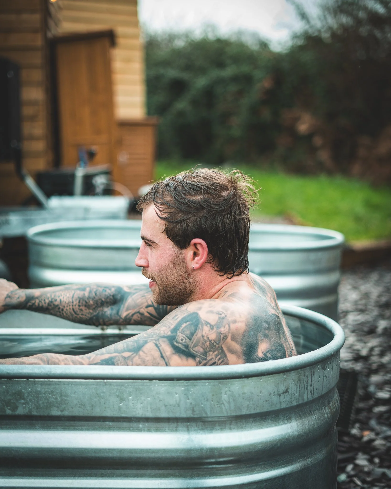 At Unfold Sauna Club Man with tattoos relaxing in an outdoor metal hot tub on a cloudy day.