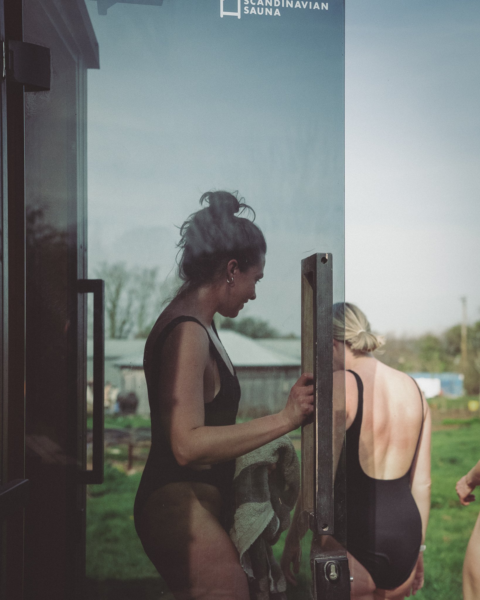At Unfold Sauna Club Two women in black swimsuits standing outside near a Scandinavian sauna, one holding a towel, with a rural landscape in the background.