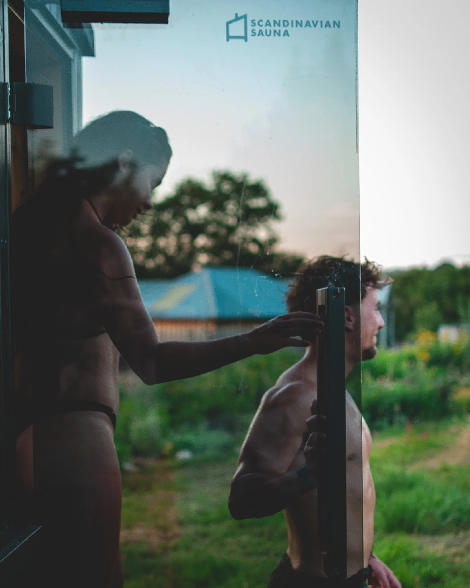 A woman with long dark hair and a man with curly hair, both shirtless, standing behind a glass door with a logo that reads 'Scandinavian Sauna'. The woman is touching the man's shoulder through the glass, and they appear to be inside a sauna or spa a