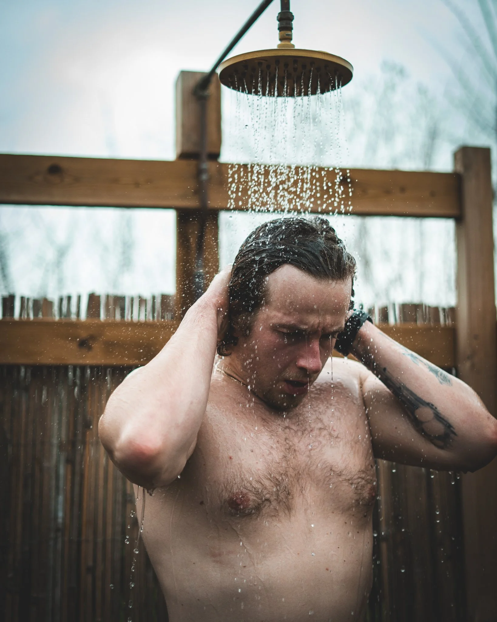 At Unfold Sauna Club Man taking outdoor shower with water pouring from overhead showerhead, during daytime, with blurred trees in background.
