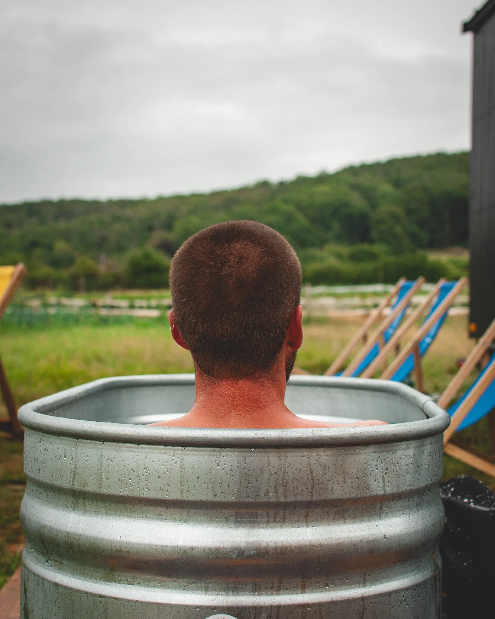 At Unfold Sauna Club Man with short hair sitting in a metal tub outdoors, with a view of green hills, cloudy sky, and deck chairs nearby.