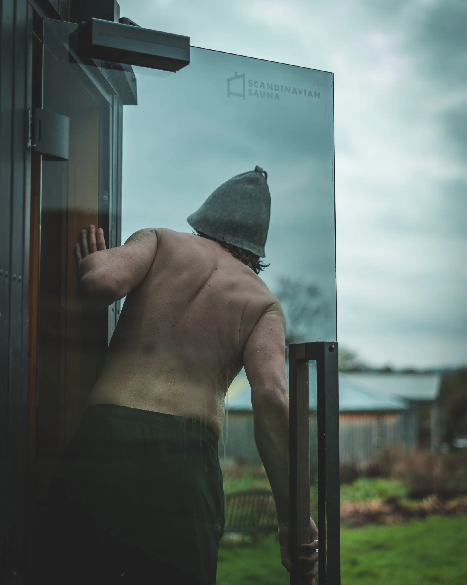 At Unfold Sauna Club A person with a grey beanie and no shirt opening a glass door outside on a cloudy day.