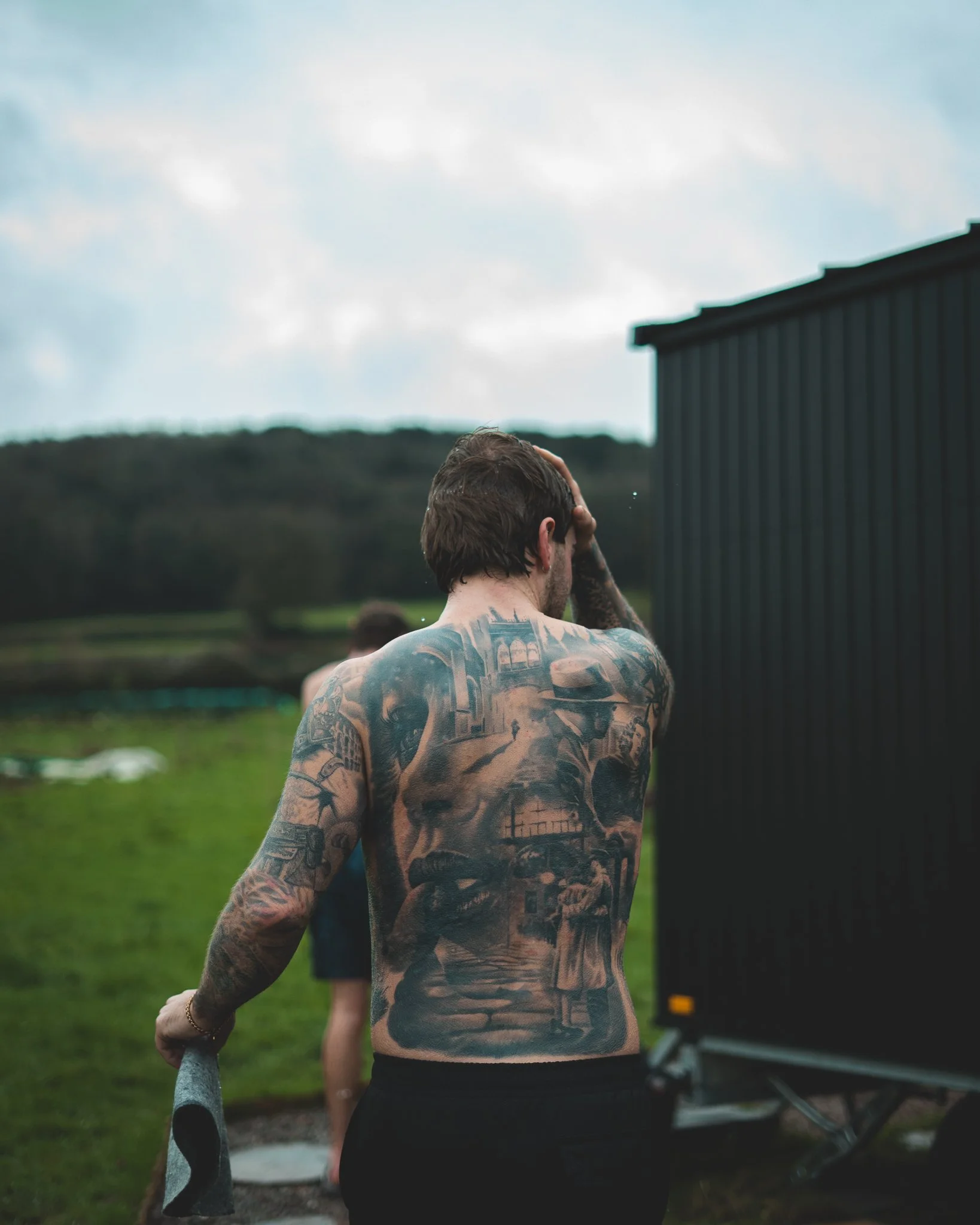 At Unfold Sauna Club Tattooed man with tattoos on his back and arms stands outdoors near a black trailer, holding a towel, with a grassy field and cloudy sky in the background.