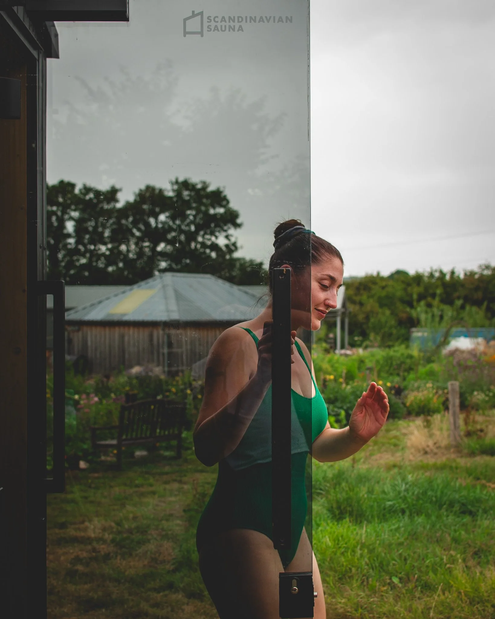 At Unfold Sauna Club A woman in a green swimsuit is seen through a glass door, enjoying a moment of outdoor relaxation in a lush garden.