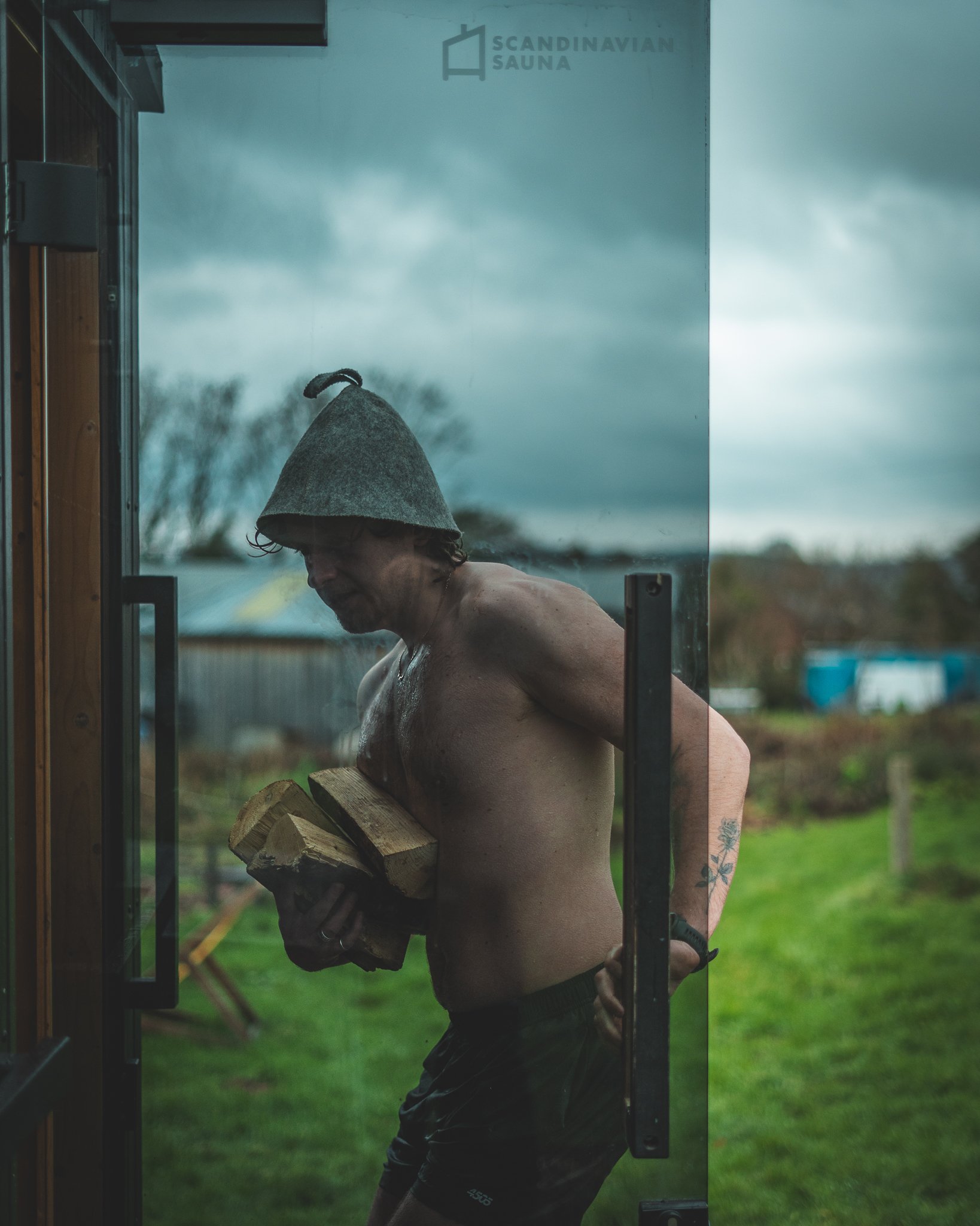At Unfold Sauna Club A topless man wearing a gray hat, holding firewood in one hand, standing outside near a glass door with a Scandinavian Sauna logo, with a cloudy sky and green field in the background.