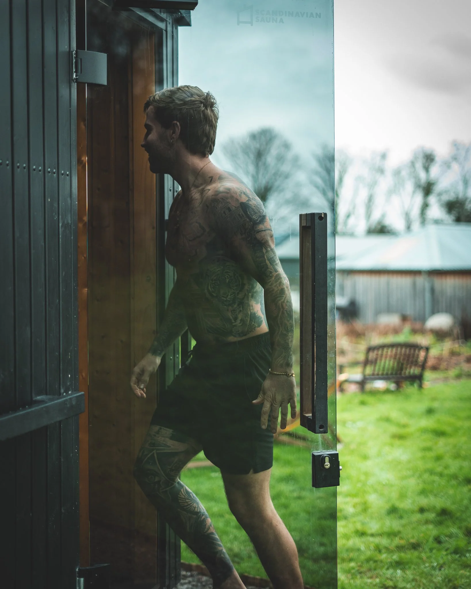 At Unfold Sauna Club A tattooed man in black shorts standing outside a sauna, looking inside, with a backyard and trees in the background.