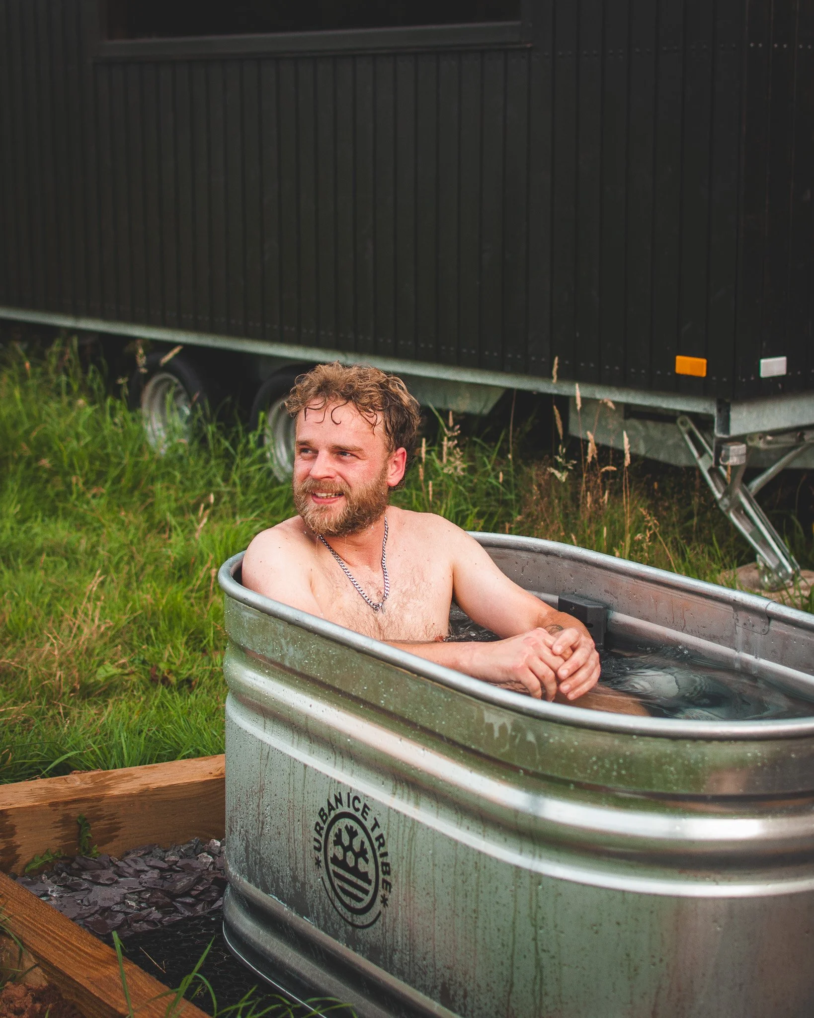 At Unfold Sauna Club A man with a beard and chain necklace sitting in a metal tub filled with water outdoors, with grass and a black trailer in the background.