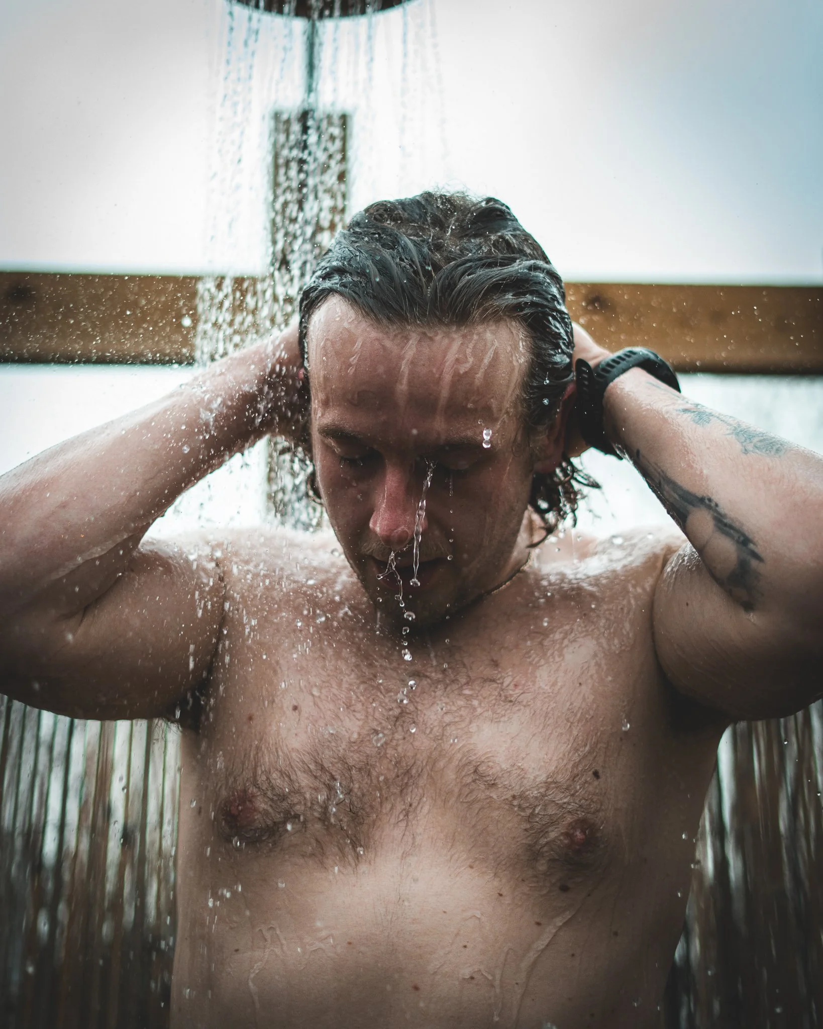 At Unfold Sauna Club A shirtless man with long hair standing under a shower with water pouring down.