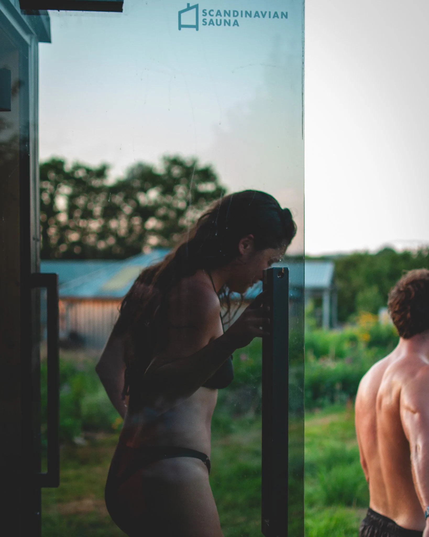 At Unfold Sauna Club A woman taking a photo outside of herself from behind a glass door at a Scandinavian sauna, with a man standing nearby in a grassy outdoor setting.