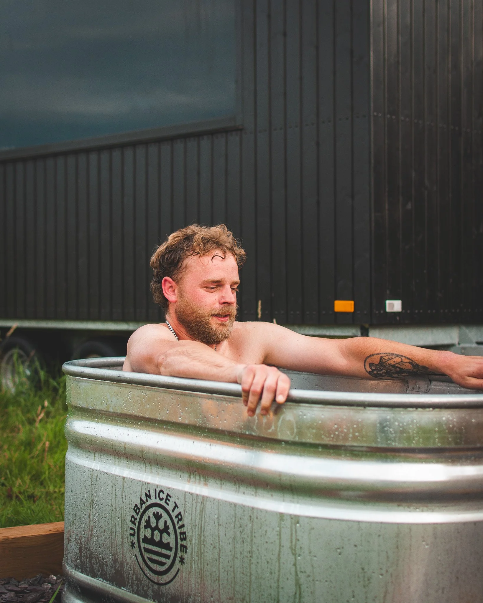 At Unfold Sauna Club A man with a beard and curly hair relaxing in an outdoor metal bathtub with the logo 'Urban Ice Trip' on it, near a black trailer, with grass and gravel nearby.