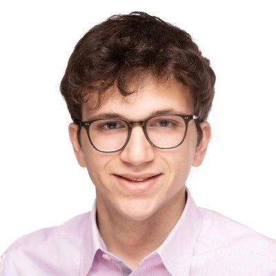 Young man with curly hair and glasses smiling in a light pink shirt against a white background.