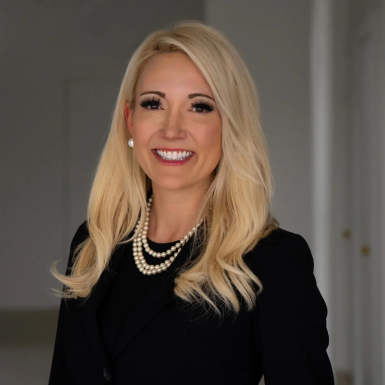 A smiling woman with long blonde hair, wearing a black blazer, double-stranded pearl necklace, and pearl earrings, standing indoors with a neutral background.