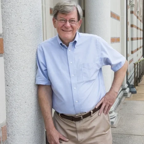 A smiling older man in a light blue button-up shirt and khaki pants leaning against a textured wall outside a building.