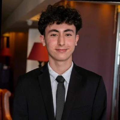 A young man with curly dark hair wearing a black suit, white shirt, and gray tie, smiling in an indoor setting with warm lighting.