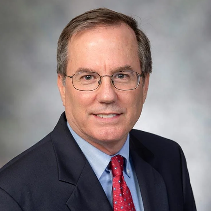 A middle-aged man wearing glasses, a dark suit, a light blue shirt, and a red tie, smiling in a professional studio portrait against a gray background.