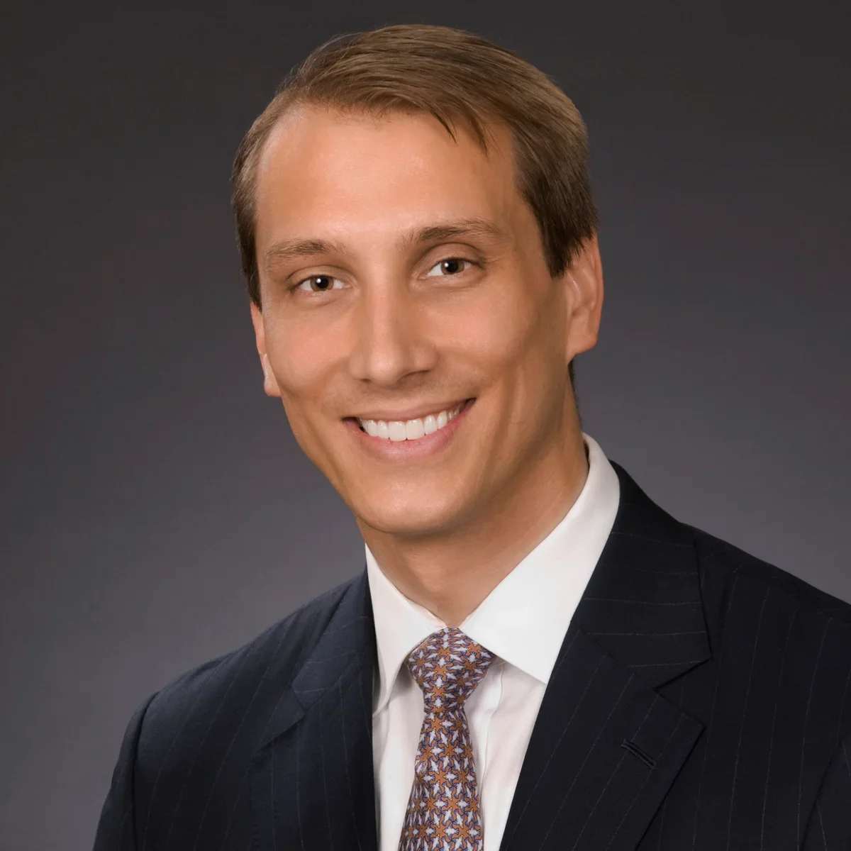 Professional headshot of a smiling man with brown hair, wearing a dark pinstripe suit, white shirt, and patterned tie against a gray background.