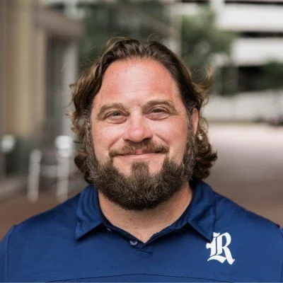 A man with curly hair and a beard smiling, wearing a blue polo shirt with a white logo on the chest.