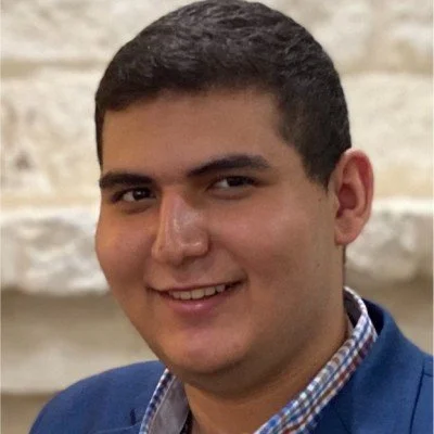 Close-up of a smiling young man with short dark hair, wearing a blue blazer and checkered shirt, standing in front of a stone wall.