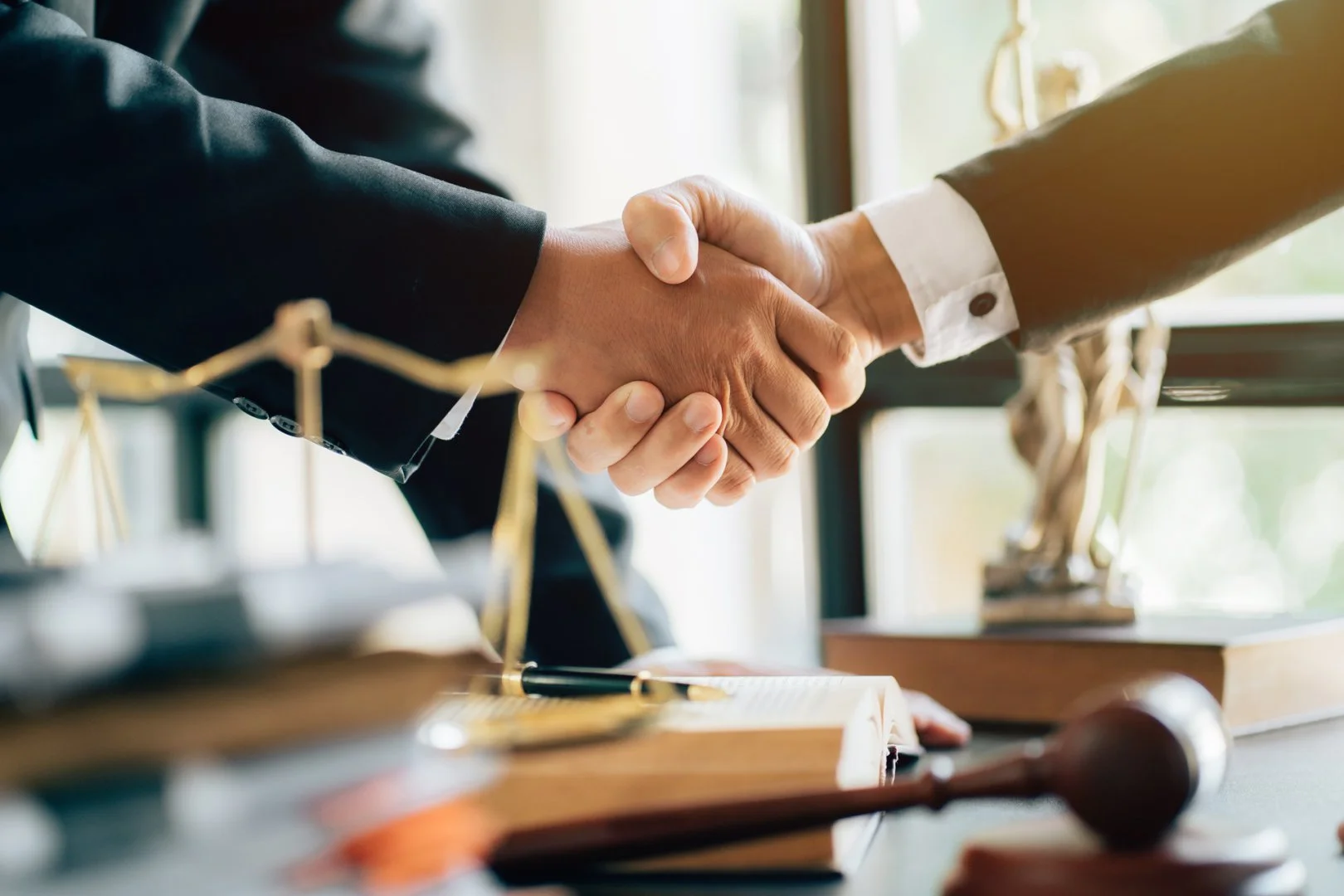 Two people in business attire shaking hands in an office setting with legal scales and gavel on the desk.