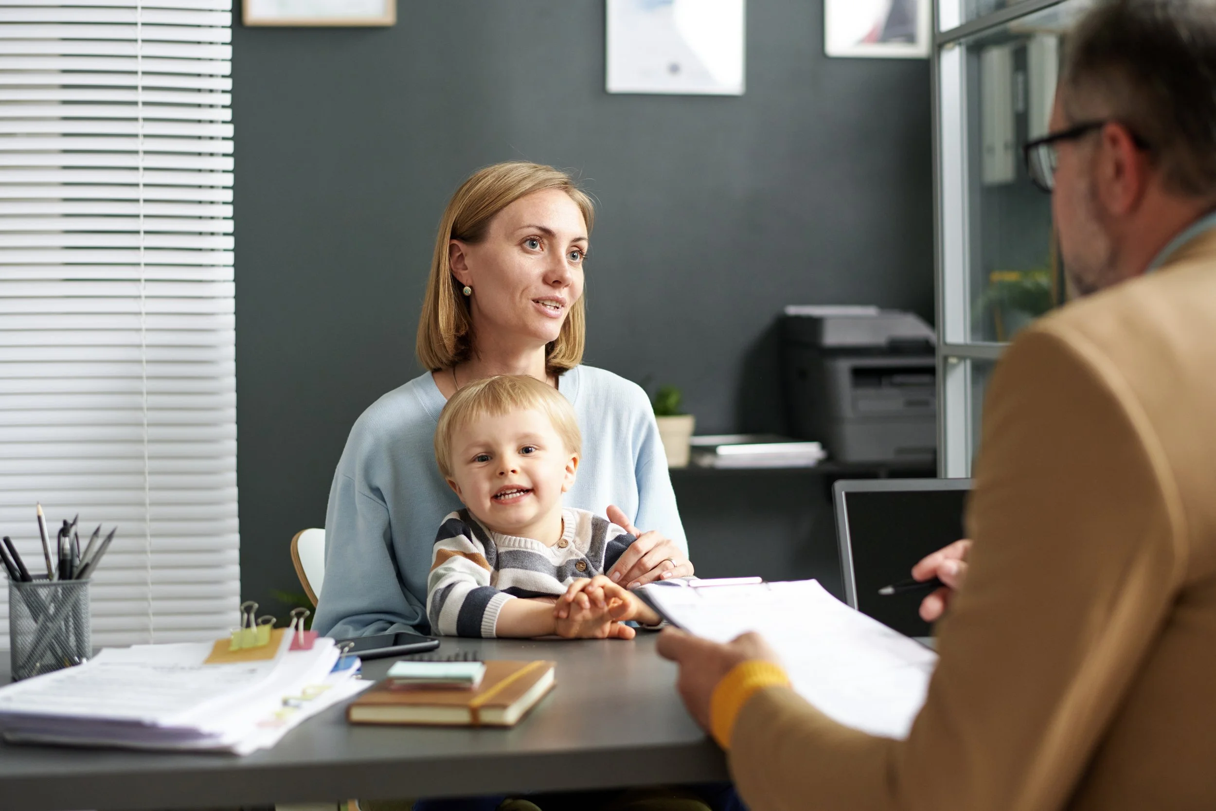 A woman with a child sitting at a desk being interviewed by a man in an office.