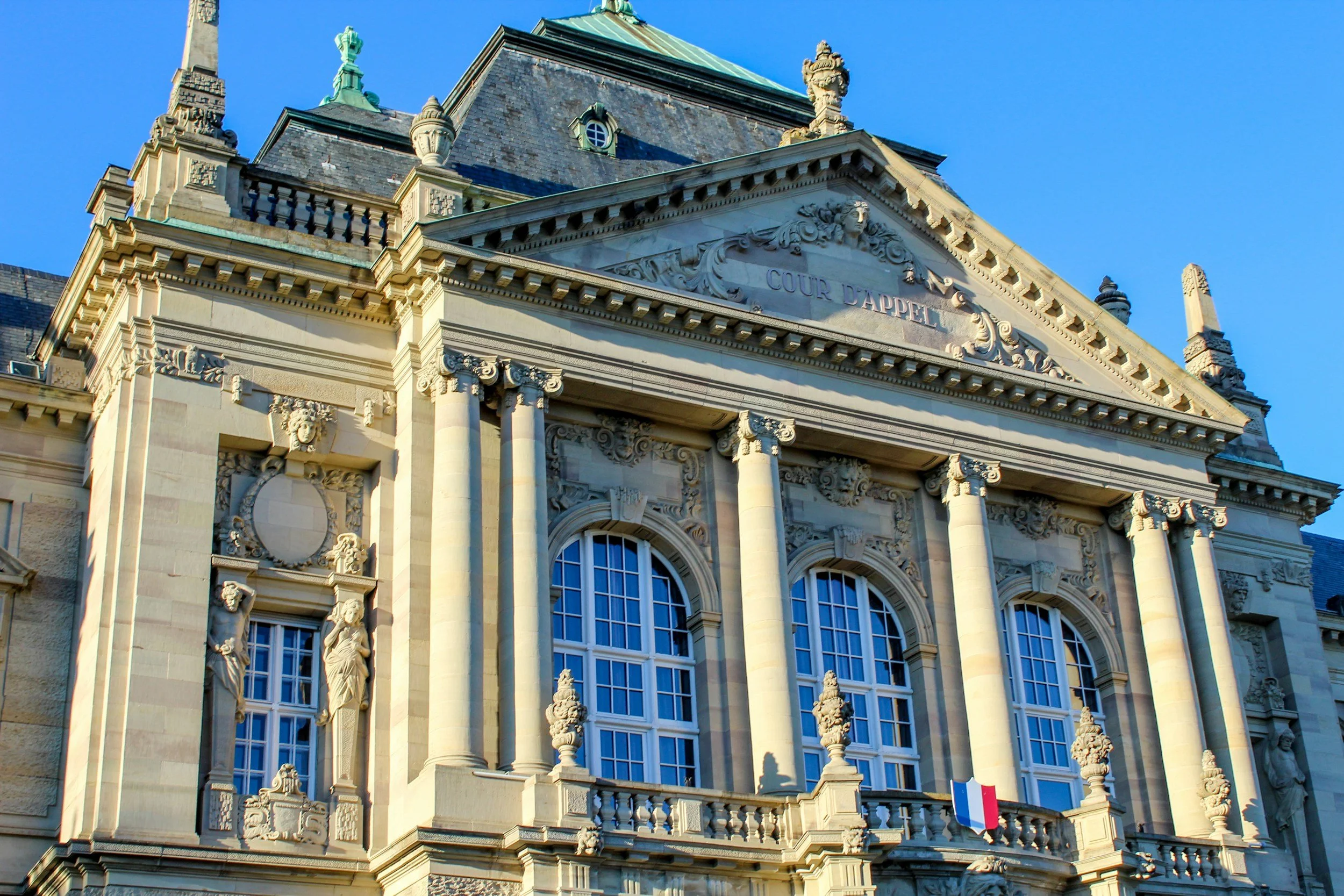 Facade of an ornate historic courthouse with intricate architectural details, large windows, sculptures, and a French flag at the bottom. The building is made of stone and features classical design elements.