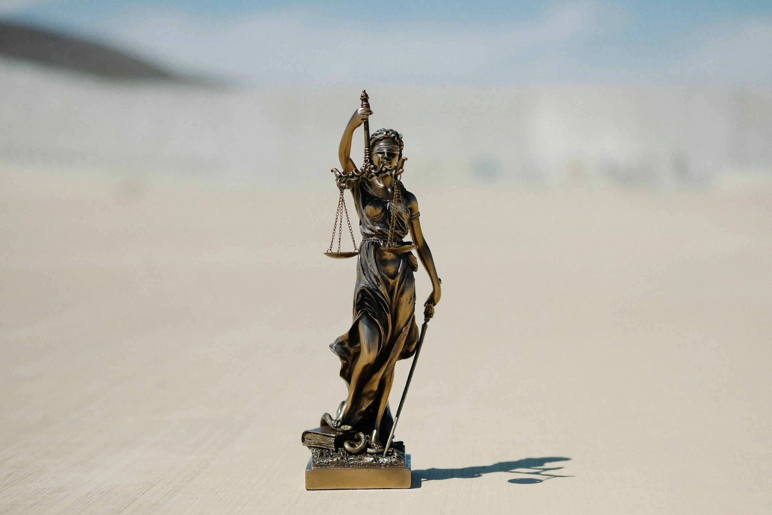 A bronze statue of Lady Justice holding scales and a sword, standing on a small rectangular base, placed on a light-colored surface outdoors under a blue sky.