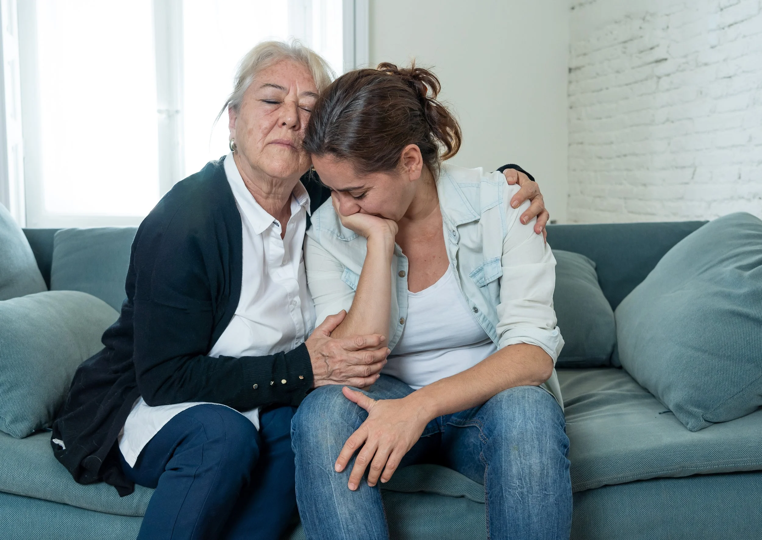 An elderly woman and a younger woman sit on a couch, embracing; the younger woman appears distressed with her face in her hand, while the elderly woman provides comfort and support.