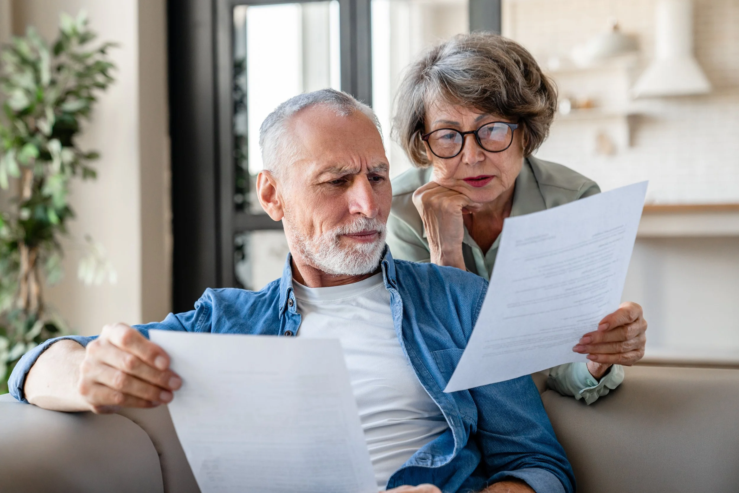 An elderly man and woman sitting on a sofa inside a home, looking at and discussing documents together. The man is holding a paper, and the woman is leaning over, examining the sheets.