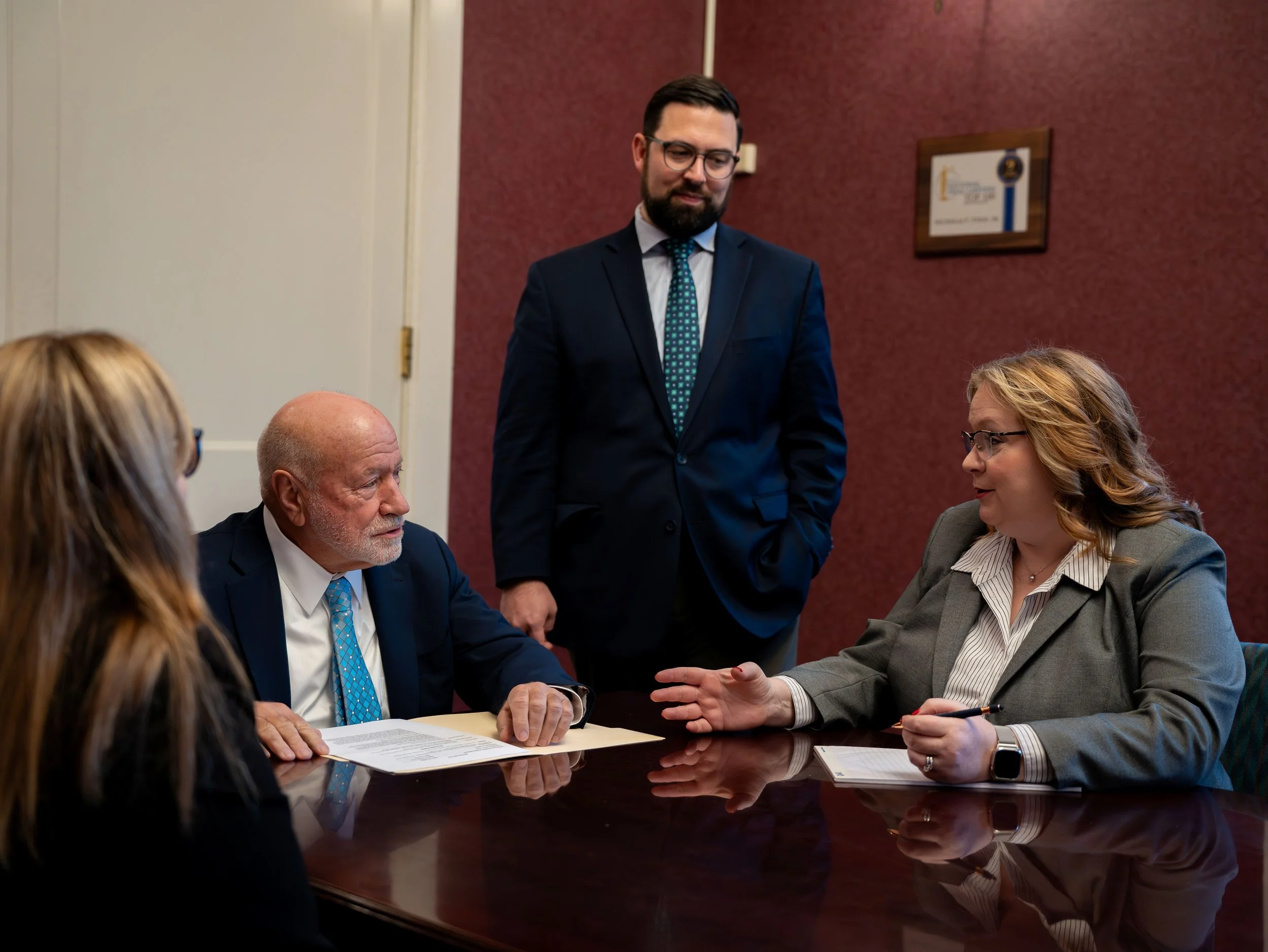 Four business professionals in a meeting room, two women and two men, engaging in a discussion around a dark wood conference table. One woman is speaking, and the others are listening attentively.