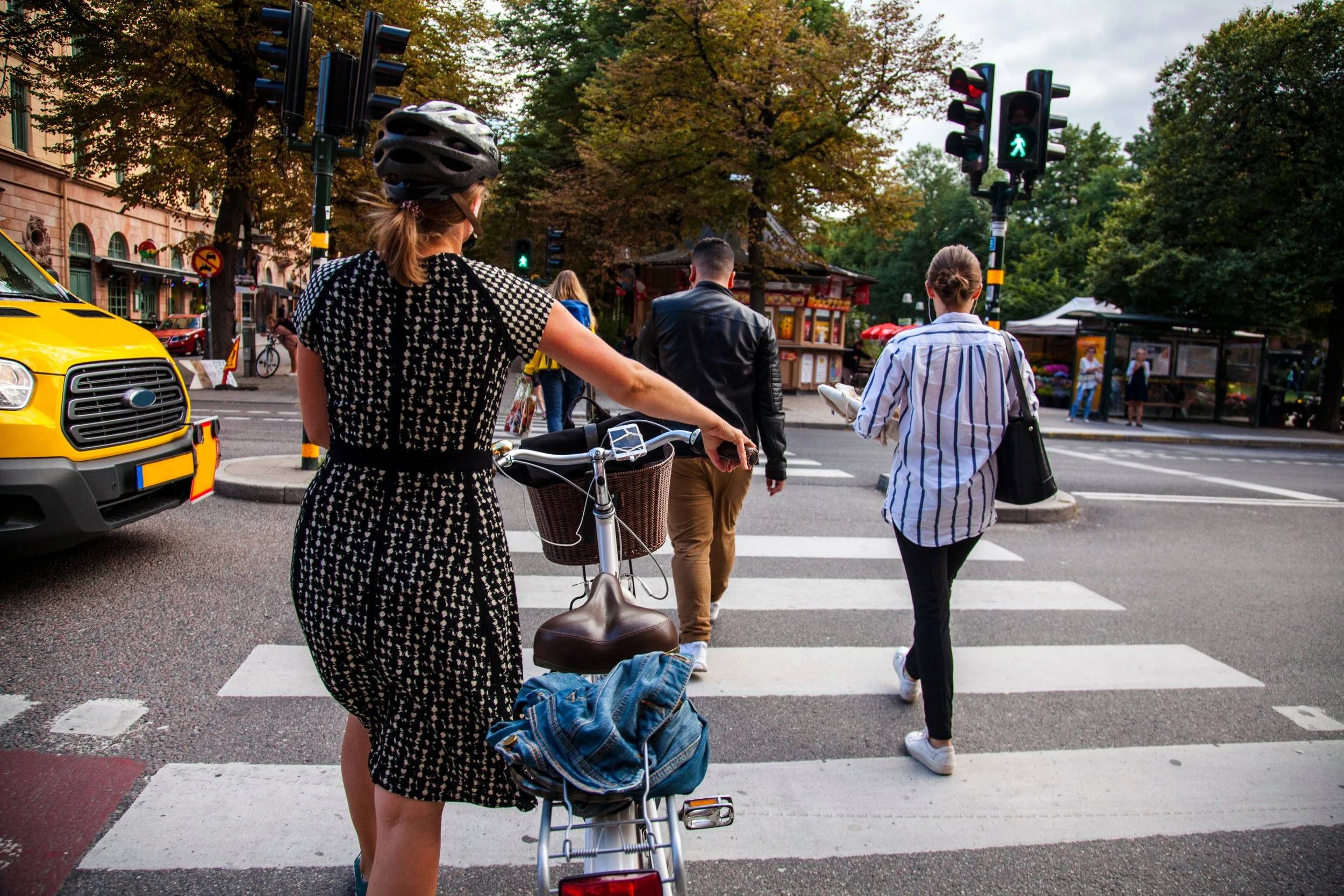 Several pedestrians crossing a city street at a crosswalk, including a woman wearing a black and white dress with a bicycle, with a basket and denim jeans, and a woman in a striped shirt carrying a black bag. There is also a man in a black jacket walking ahead.